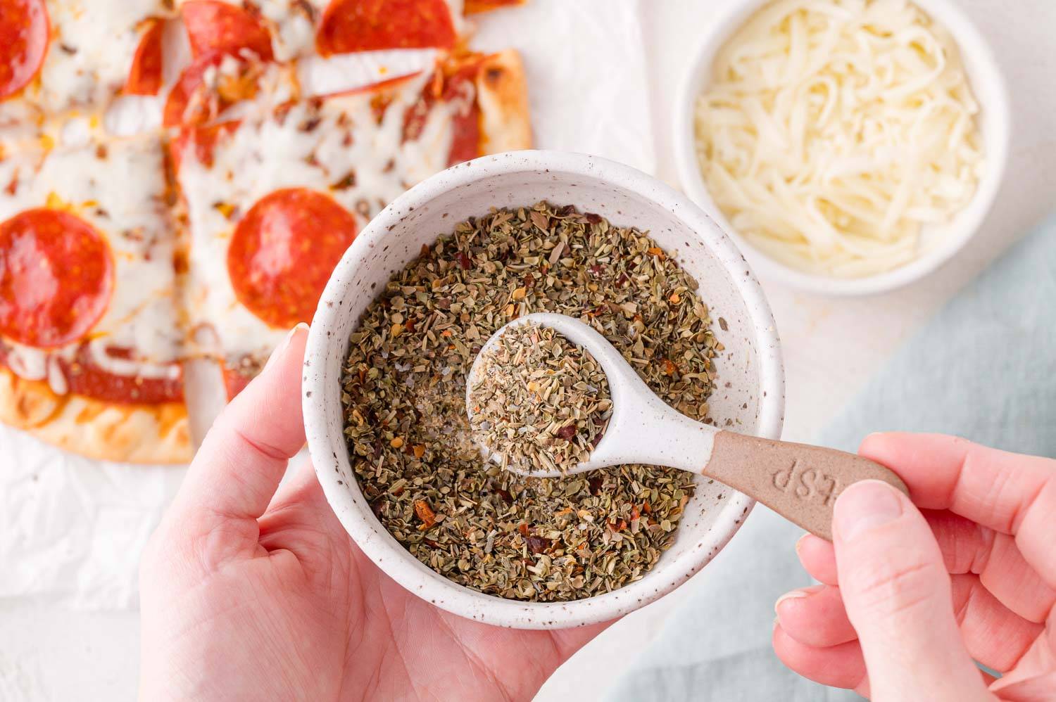 Pizza seasoning in a small bowl, with a measuring spoon held in a hand. Both are held over a pepperoni pizza.