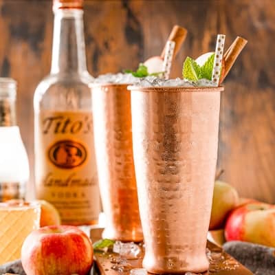 Two copper cups with paper straws, and ice showing. A bottle of titos vodka in the background.