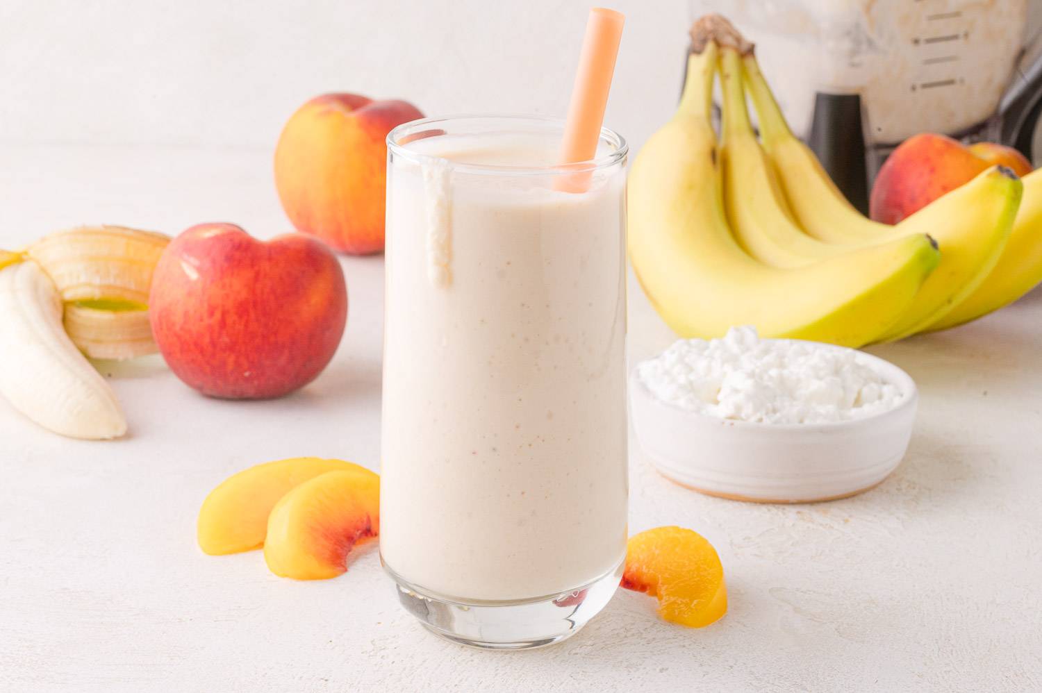 Cottage cheese smoothie in a glass with orange straw, peaches and bananas in background.