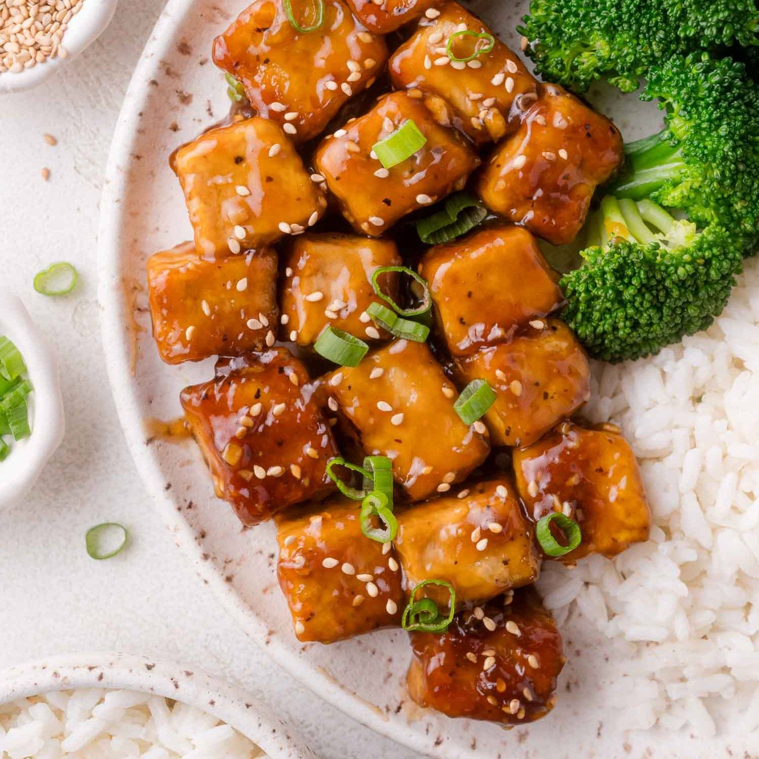 Glossy teriyaki glazed tofu cubes on a plate with broccoli and rice.