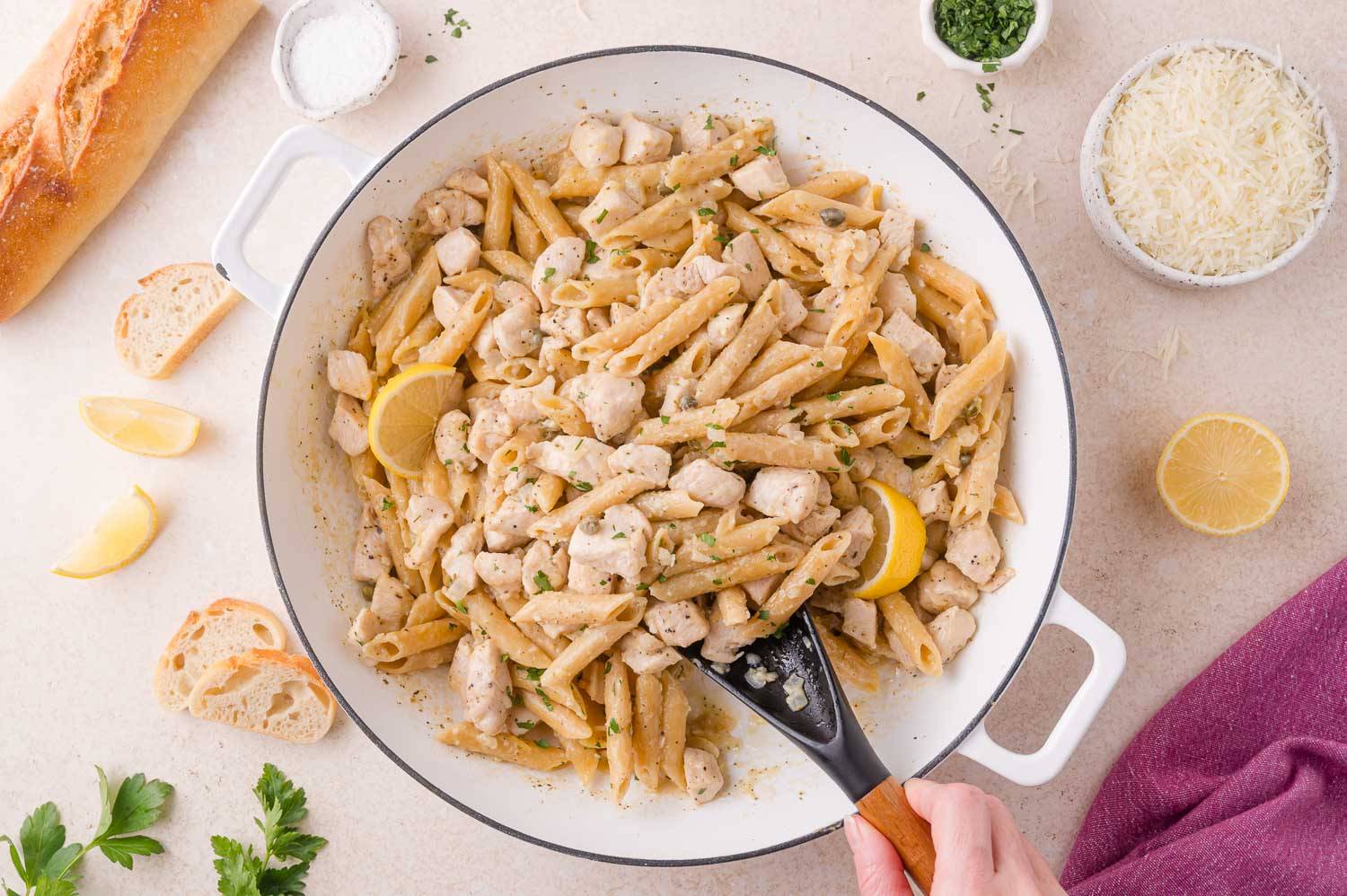 Chicken piccata pasta in a pan, penne pasta, chicken, fresh herbs, and a creamy sauce.