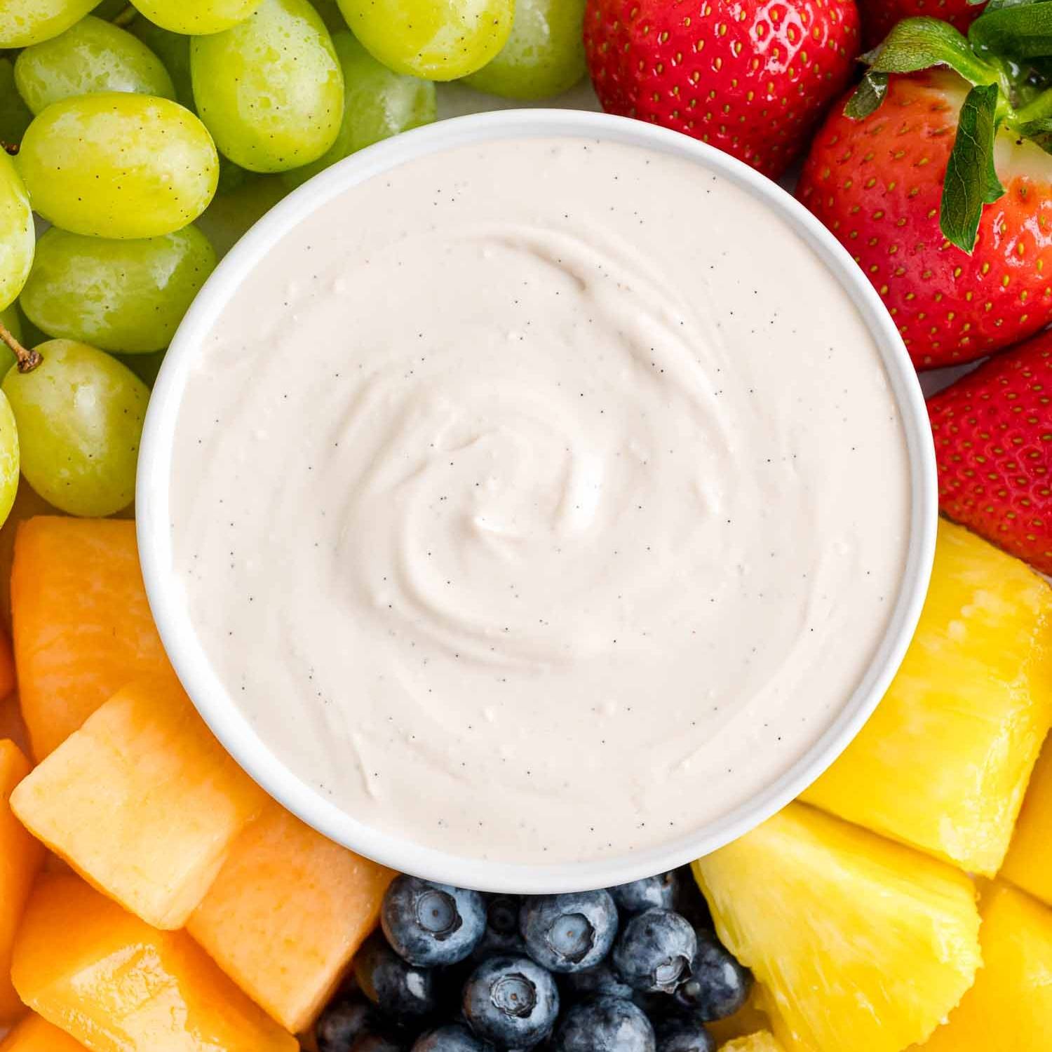 Fruit dip in a round bowl, surrounded by grapes, strawberries, pineapple, blueberries, and melon.