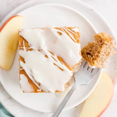 Slice of apple cake with icing, on a white plate, a fork holding a small portion of it.