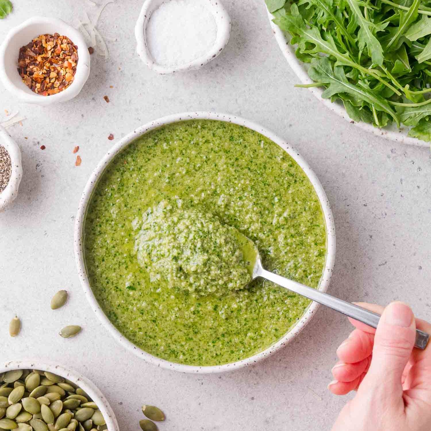 Arugula pesto in a small bowl, with a hand holding a spoon scooping some out.