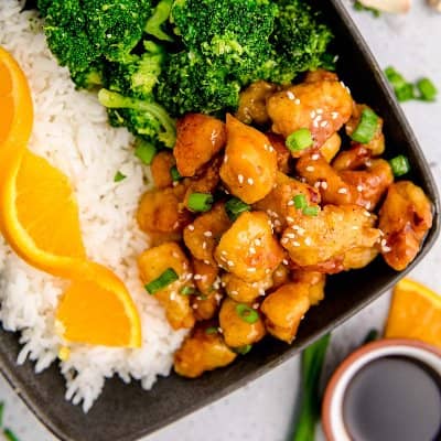 Small pieces of glazed chicken in a bowl with rice, broccoli, and topped with sesame seeds and green onions.