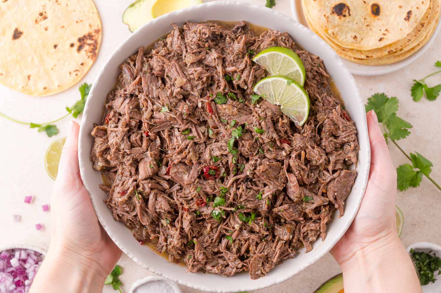 Beef barbacoa held in two hands, with tortillas, red onions, and cilantro in the background.
