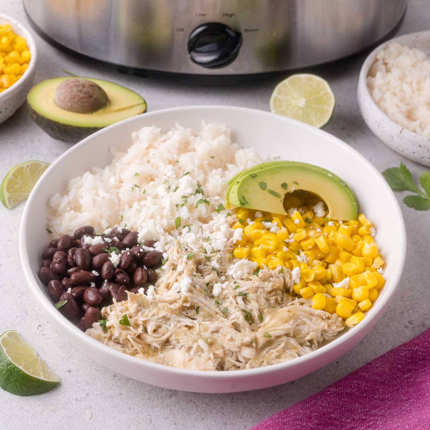 Bowl in front of a slow cooker, bowl contains shredded salsa verde chicken, corn, avocado, rice, and black beans.