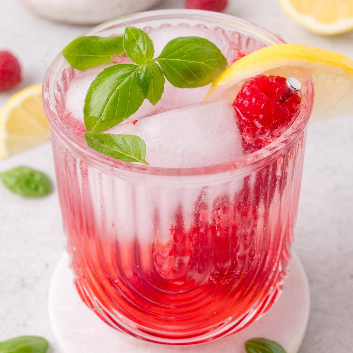 Mocktail in decorative glass, garnished with raspberries, basil, and lemon.