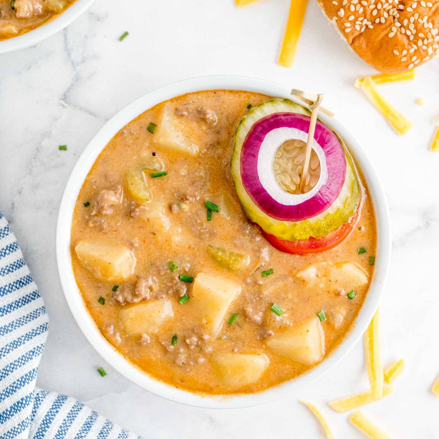 Chunky cheeseburger soup in a small white bowl, garnished with tomato, pickle, onion.