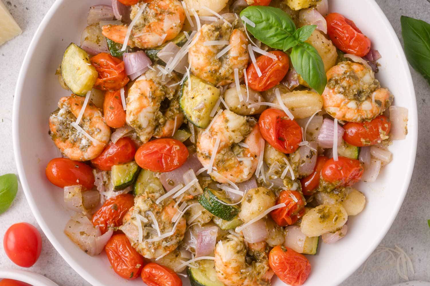 White bowl containing shrimp, vegetables, and gnocchi, all tossed in pesto and garnished with shredded parmesan cheese.