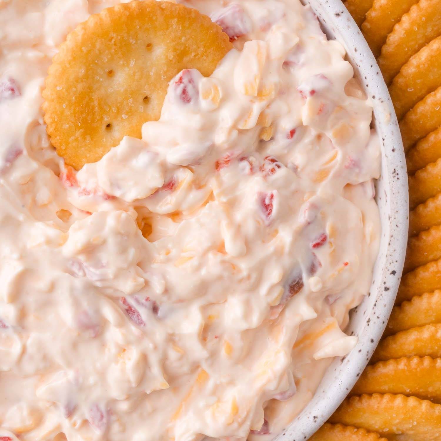 Pimento cheese in a bowl, a ritz cracker dipped in it. More ritz crackers on the right side of the bowl.