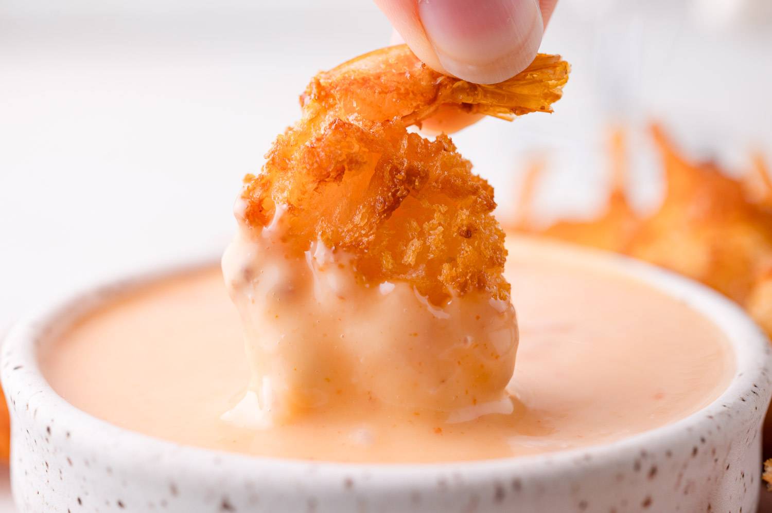 Breaded shrimp being dipped into bang bang sauce in a speckled bowl.