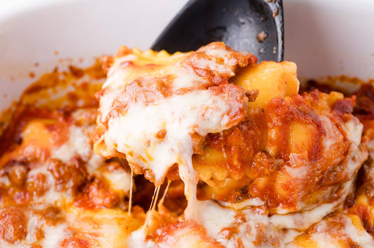 Baked ravioli being scooped out of a casserole dish with a black spoon.