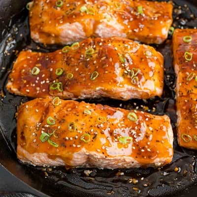 Salmon in a black skillet glazed with teriyaki sauce and garnished with green onions and sesame seeds.