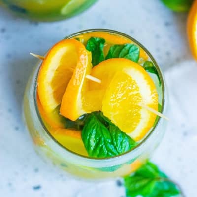 Brightly colored drink in stemless glass with a twist of orange and fresh mint leaves.