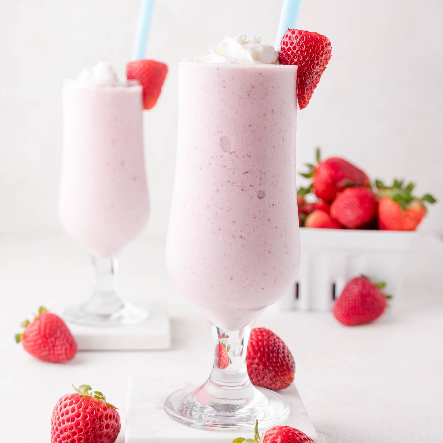 Two strawberry milkshakes in tall glasses, topped with a strawberry and whipped cream.