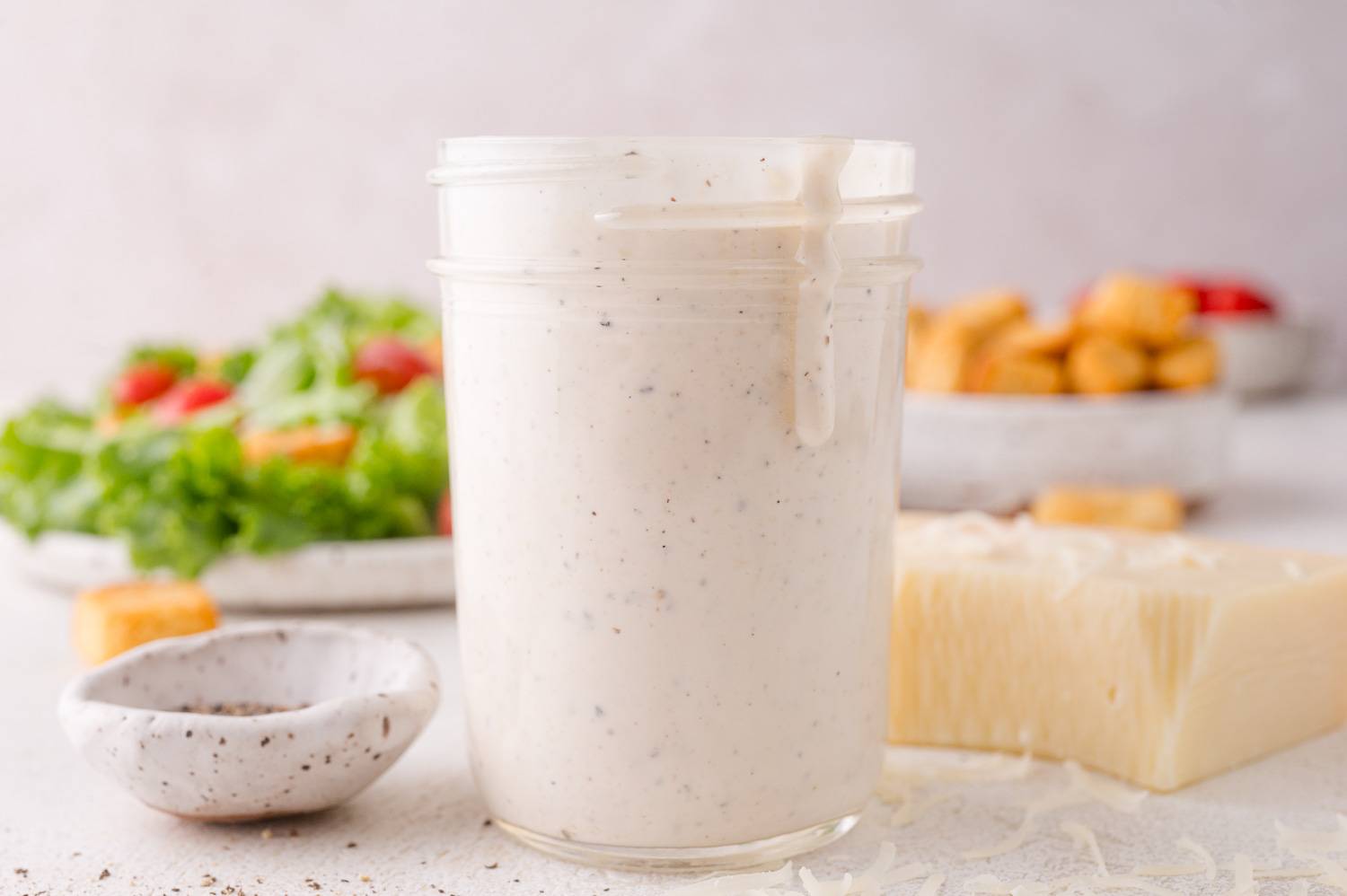 Parmesan peppercorn dressing in a small clear glass jar, salad ingredients in the background on the photo.