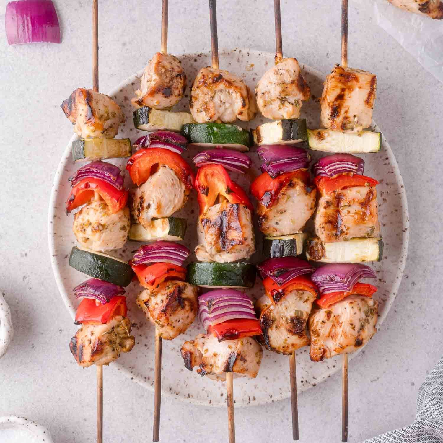 Lined up chicken kabobs on a plate with red bell peppers, zucchini, onion.