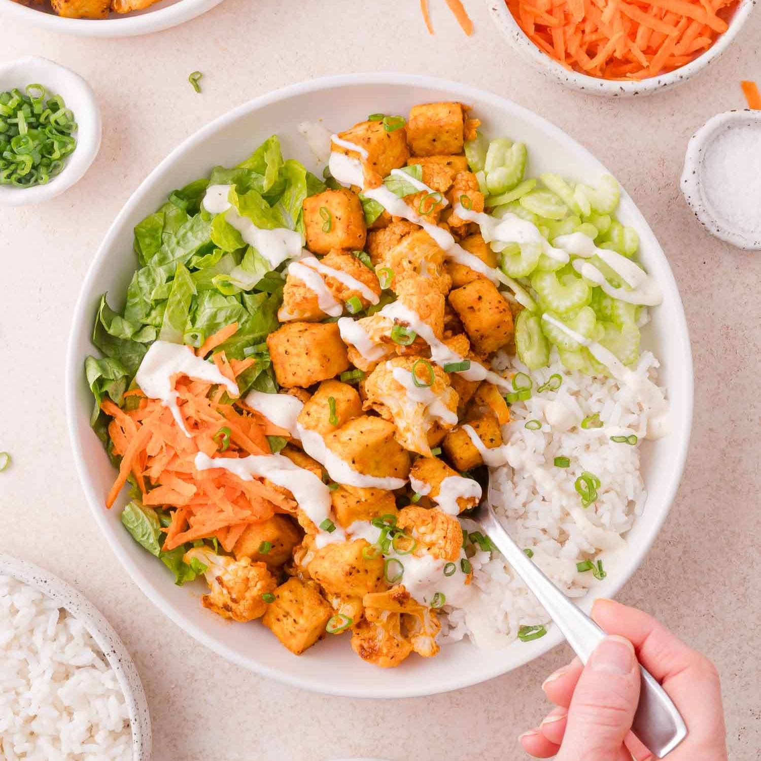 A white bowl filled with buffalo tofu, cauliflower, rice, celery, carrots, and lettuce. 