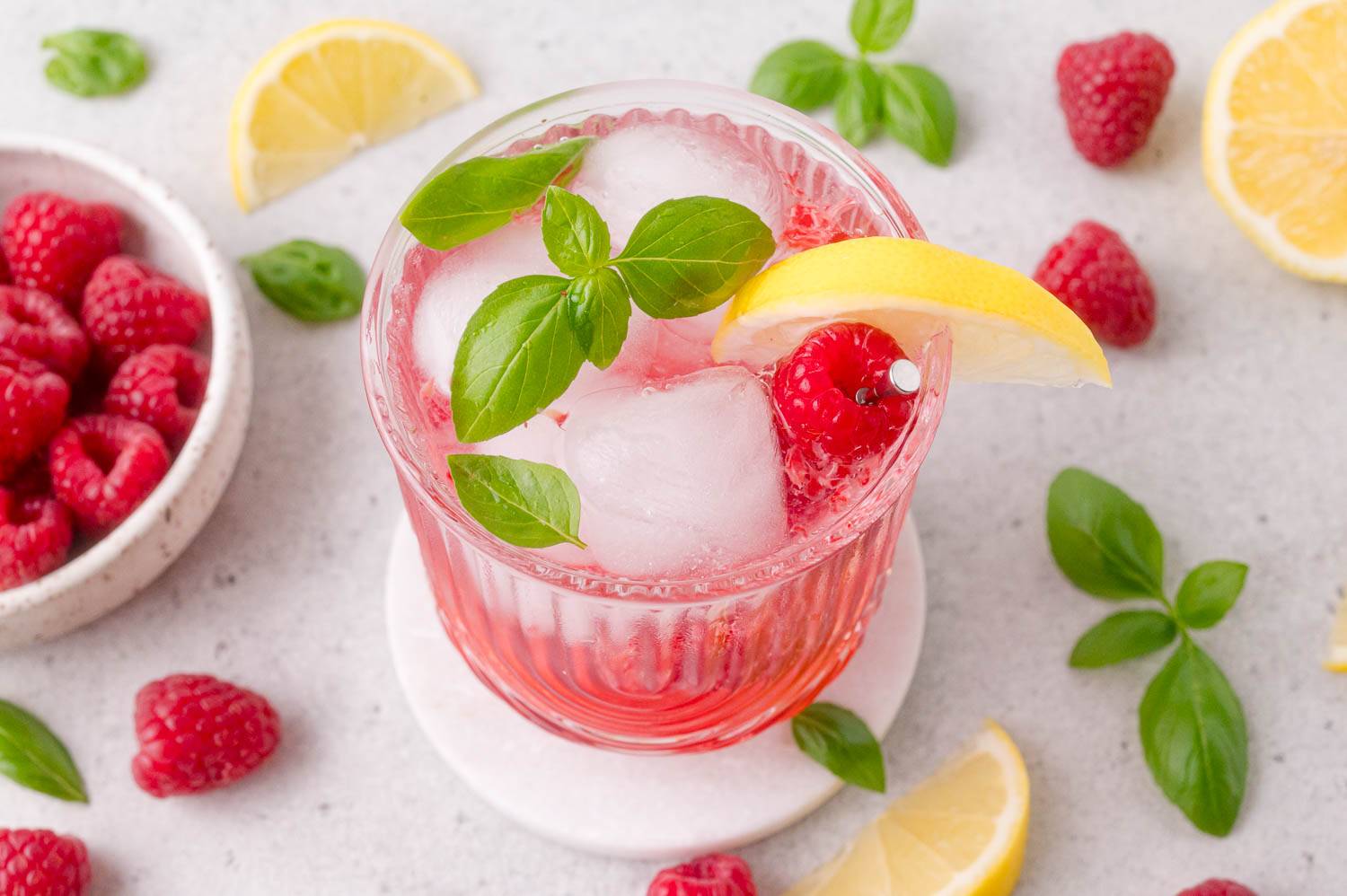Cocktail glass with a fresh raspberry drink, garnished with lemon and basil.