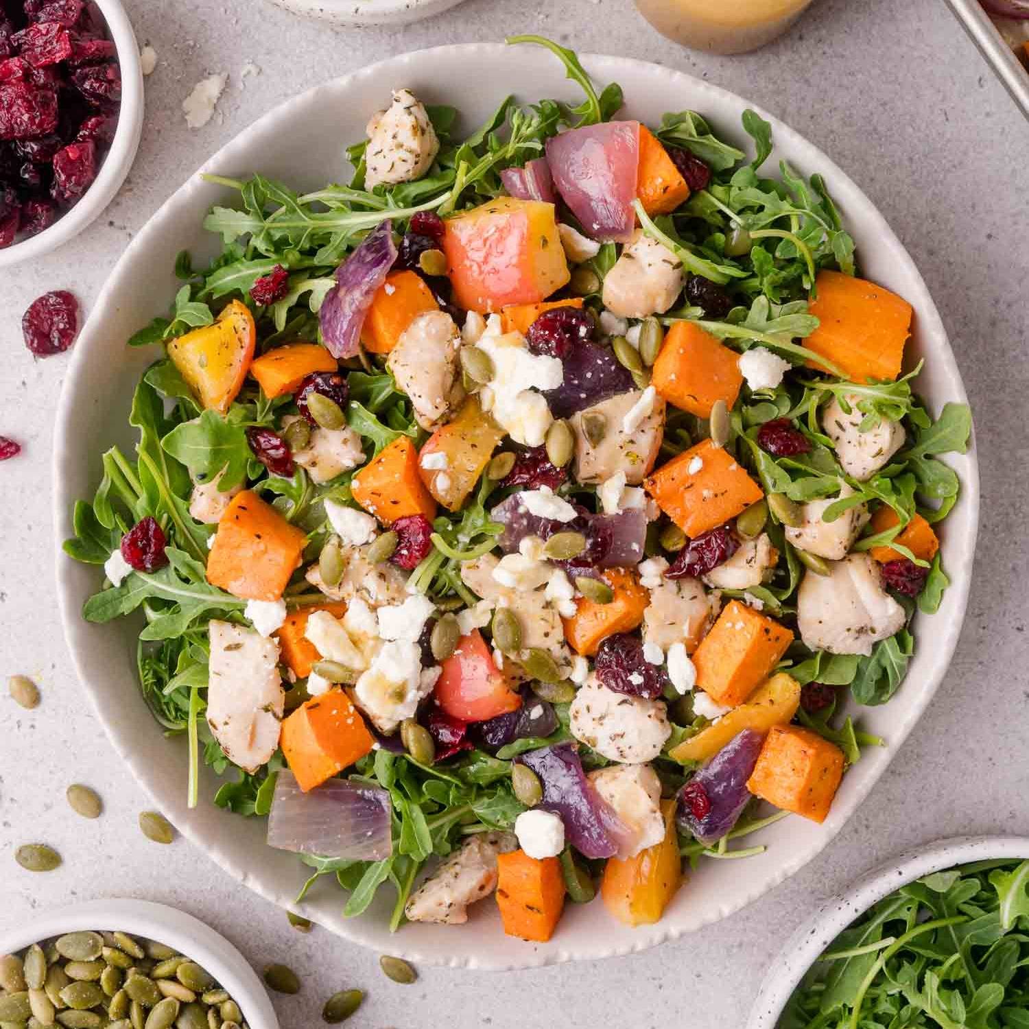 A salad with arugula, roasted sweet potatoes, onions, chicken, apples, garnished with pepitas and feta.