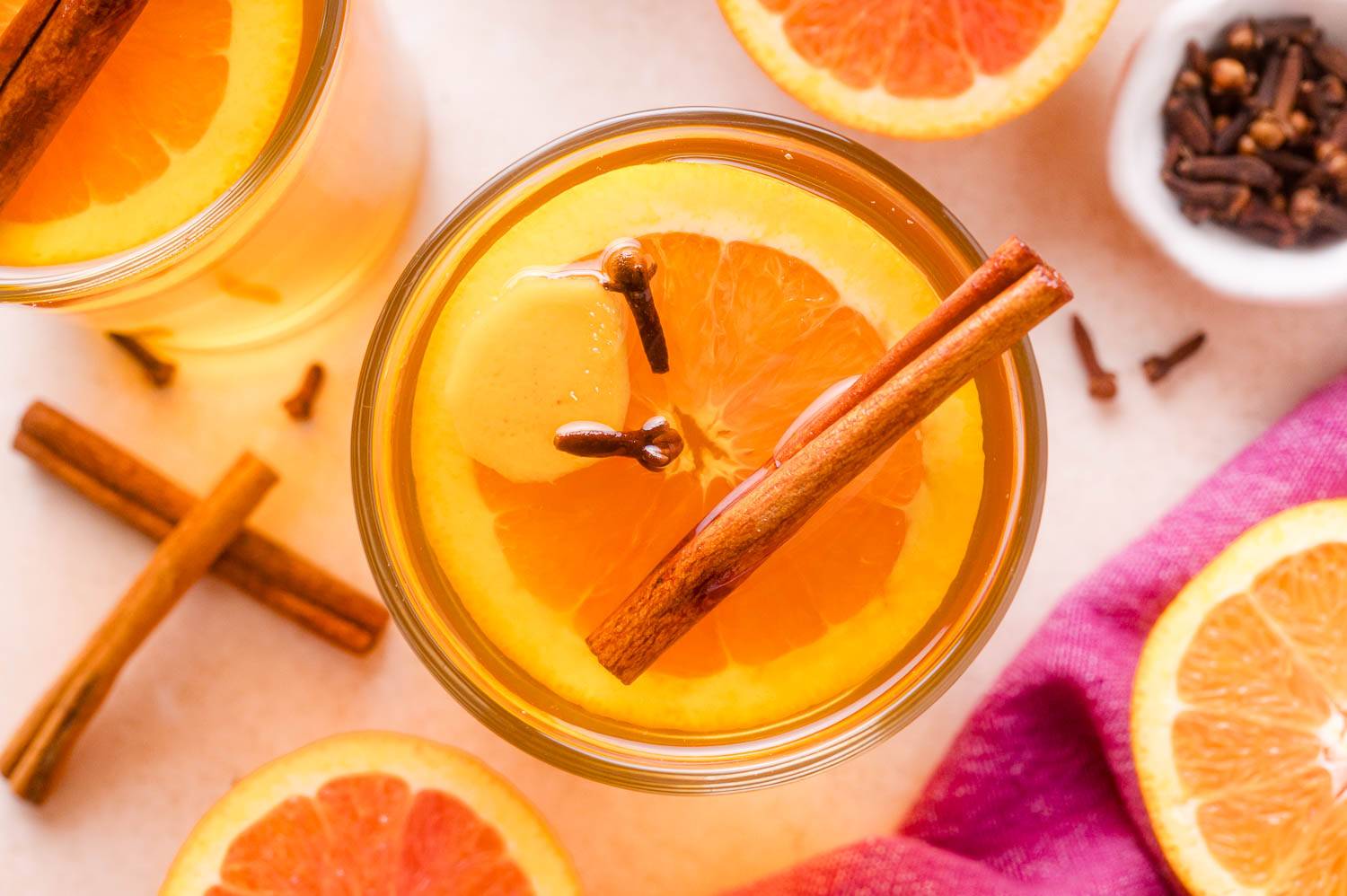 Mulled apple cider in a glass with a slice of ginger, an orange slice, a cinnamon stick, and a couple of cloves.