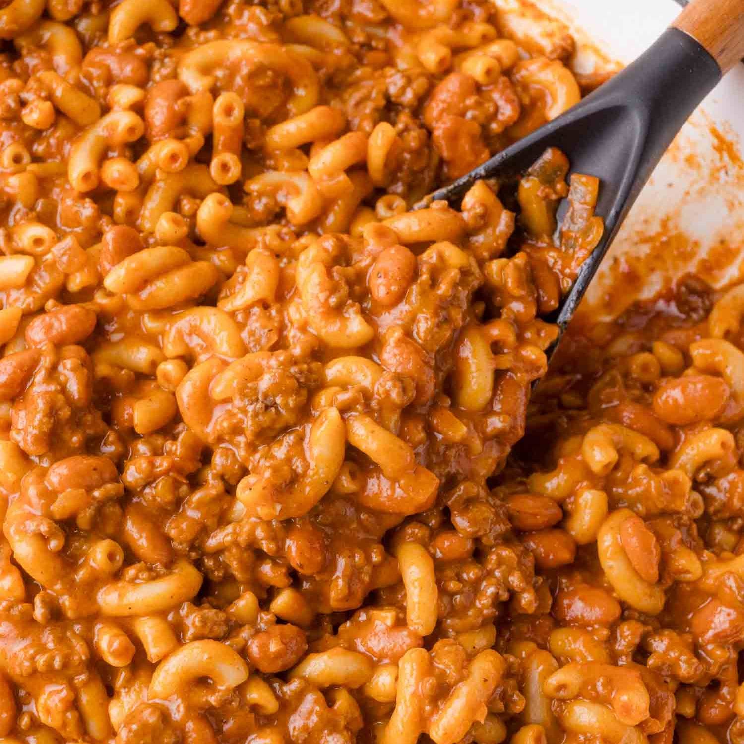 Macaroni noodles, ground beef, and pinto beans in a creamy tomato sauce with a black spoon.