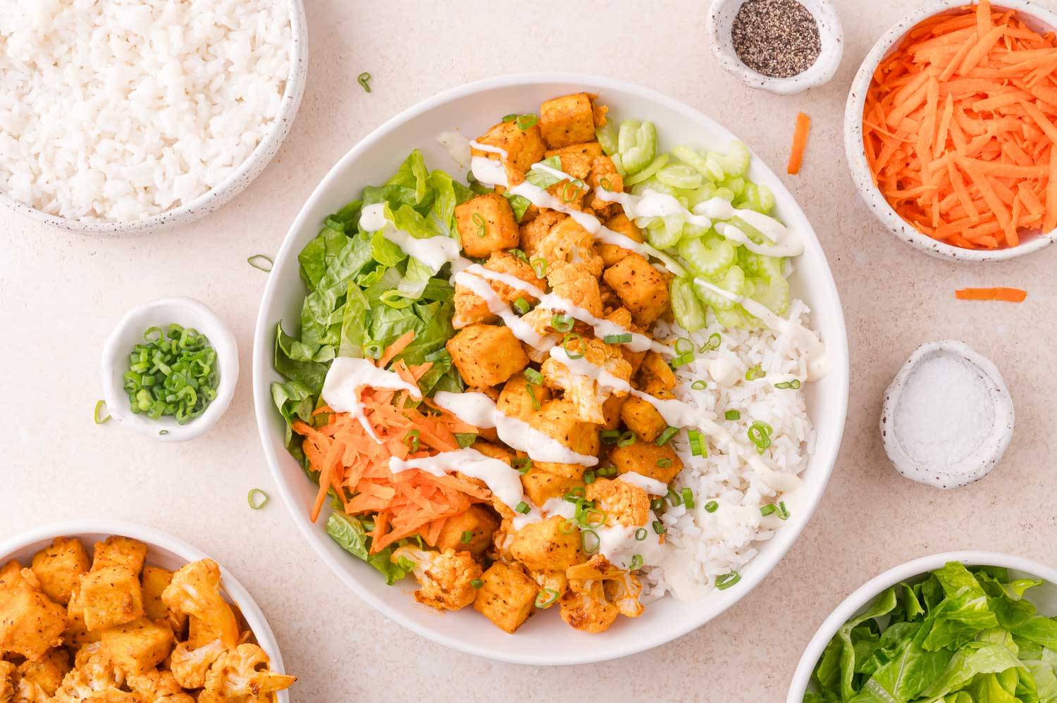 Buffalo chicken and cauliflower in a bowl with rice, lettuce, celery, carrots, and ranch drizzle.