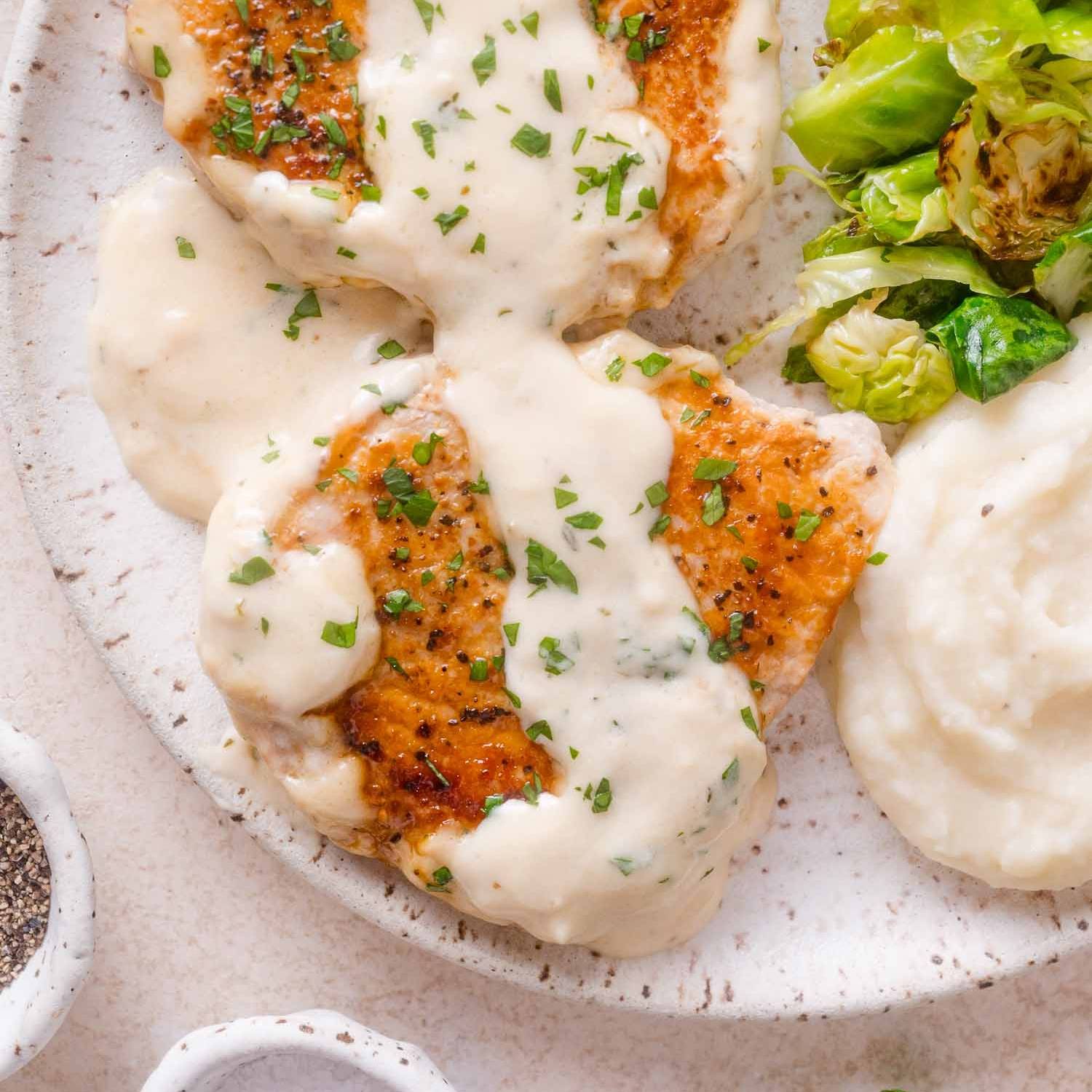 Pork chops with a creamy white sauce on top, on a plate with mashed potatoes and Brussels sprouts.