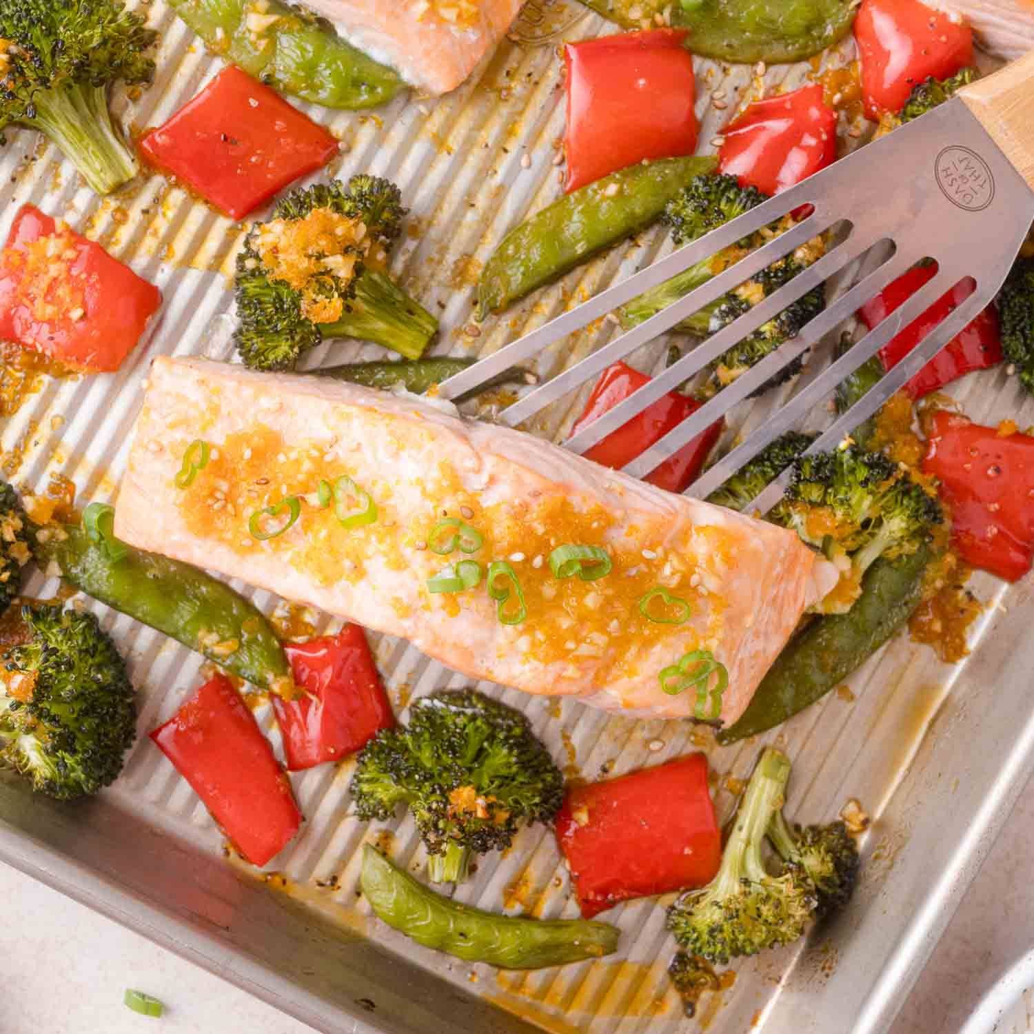 Salmon on a sheet pan with roasted vegetables, being lifted with a spatula.