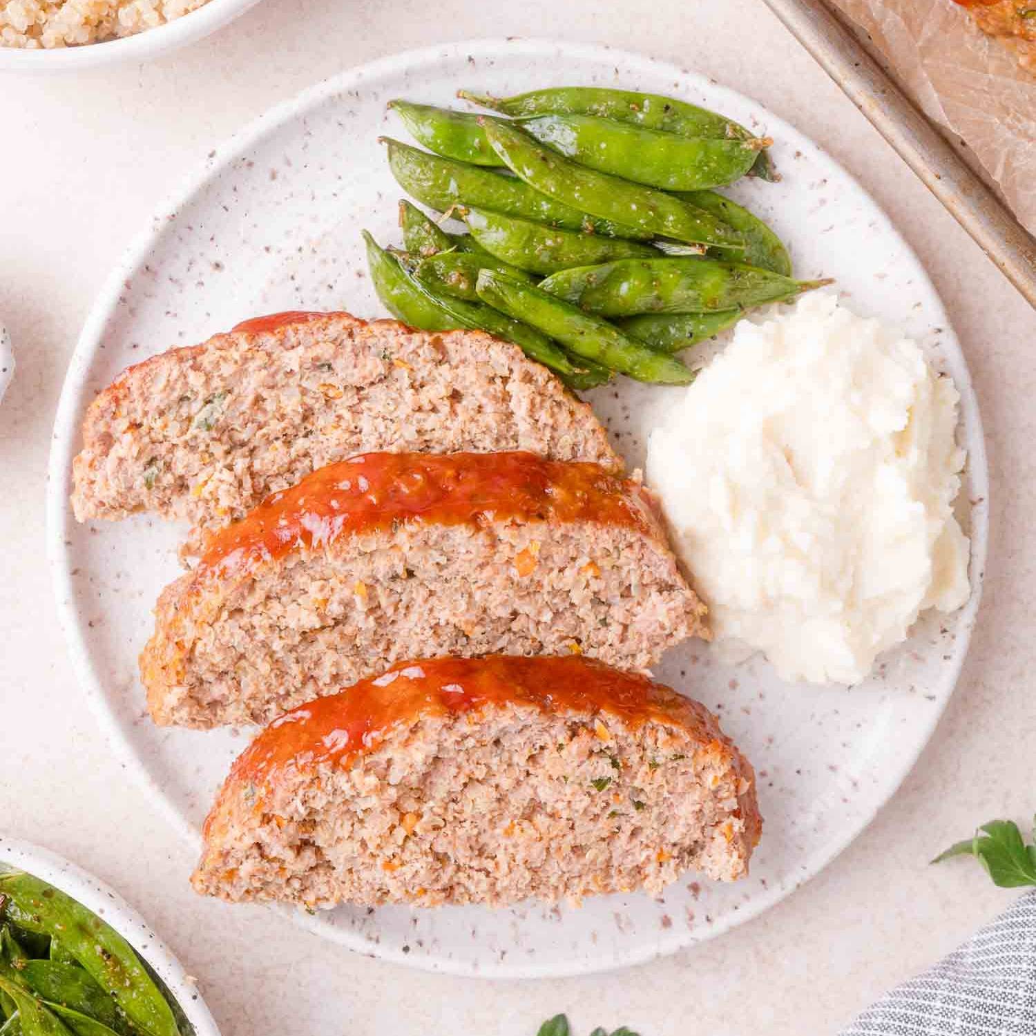 Three slices of turkey meatloaf on a plate with snap peas and mashed potatoes.