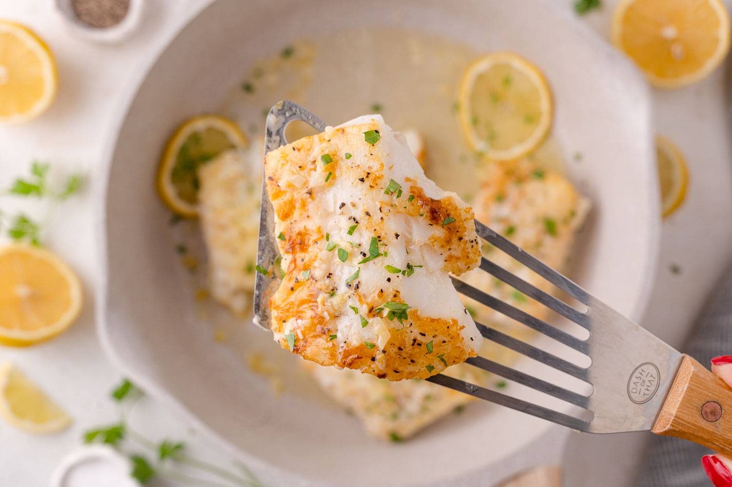 Lemon butter cod being lifted out of a pan with. more cod fillets.