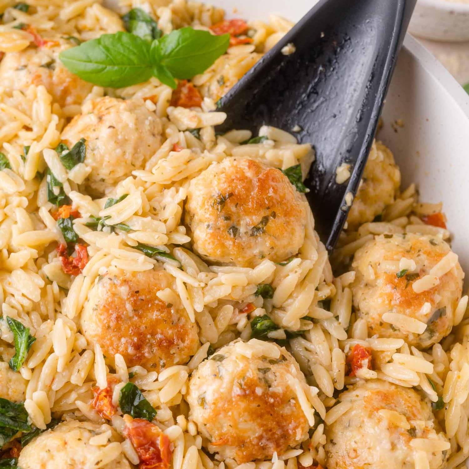 Meatballs, sundried tomatoes, and spinach in a white skillet with a black spoon.