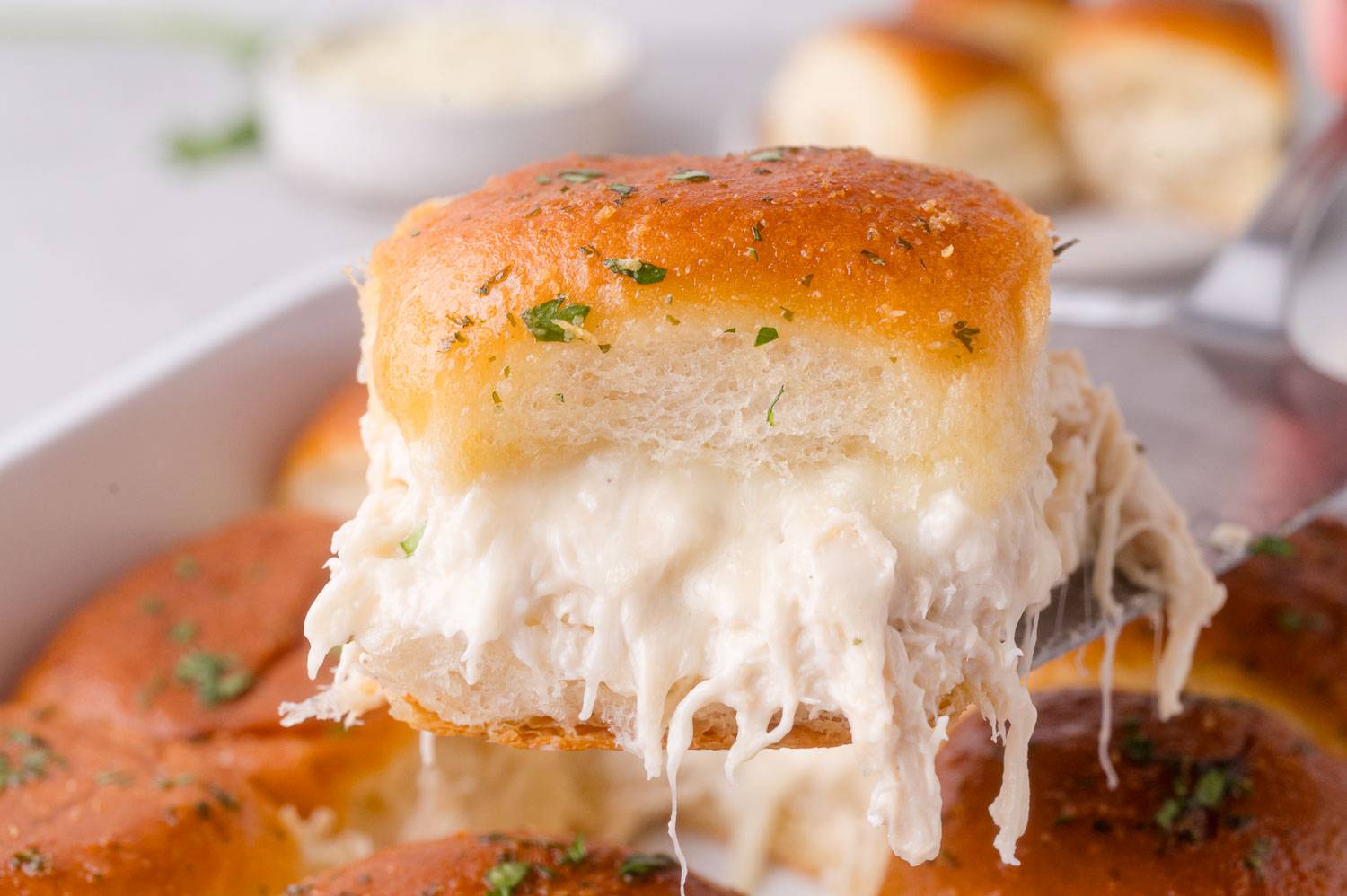 Cheesy chicken slider being lifted out of a pan containing more sliders.