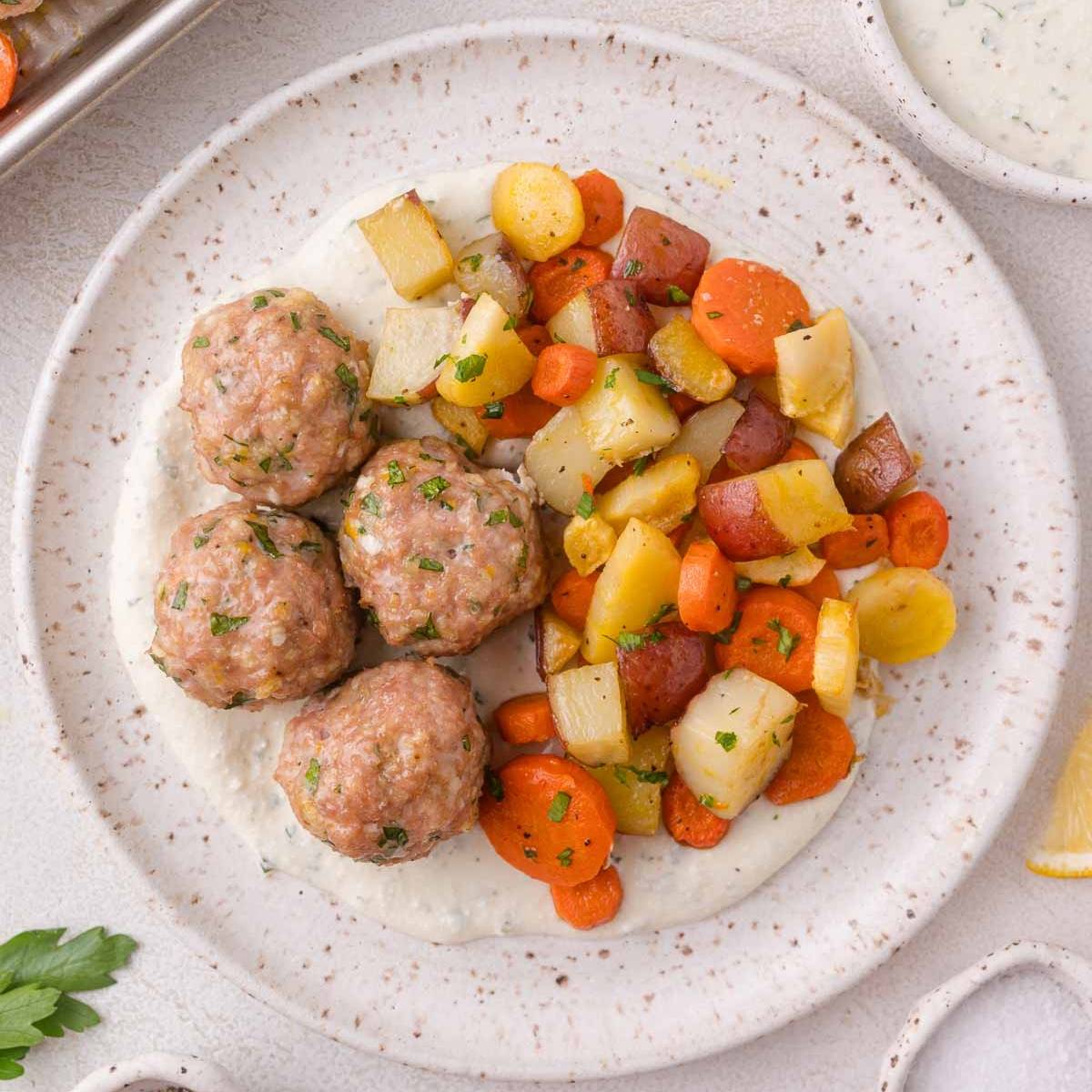 Meatballs on a plate with colorful roasted root vegetables and a white sauce.