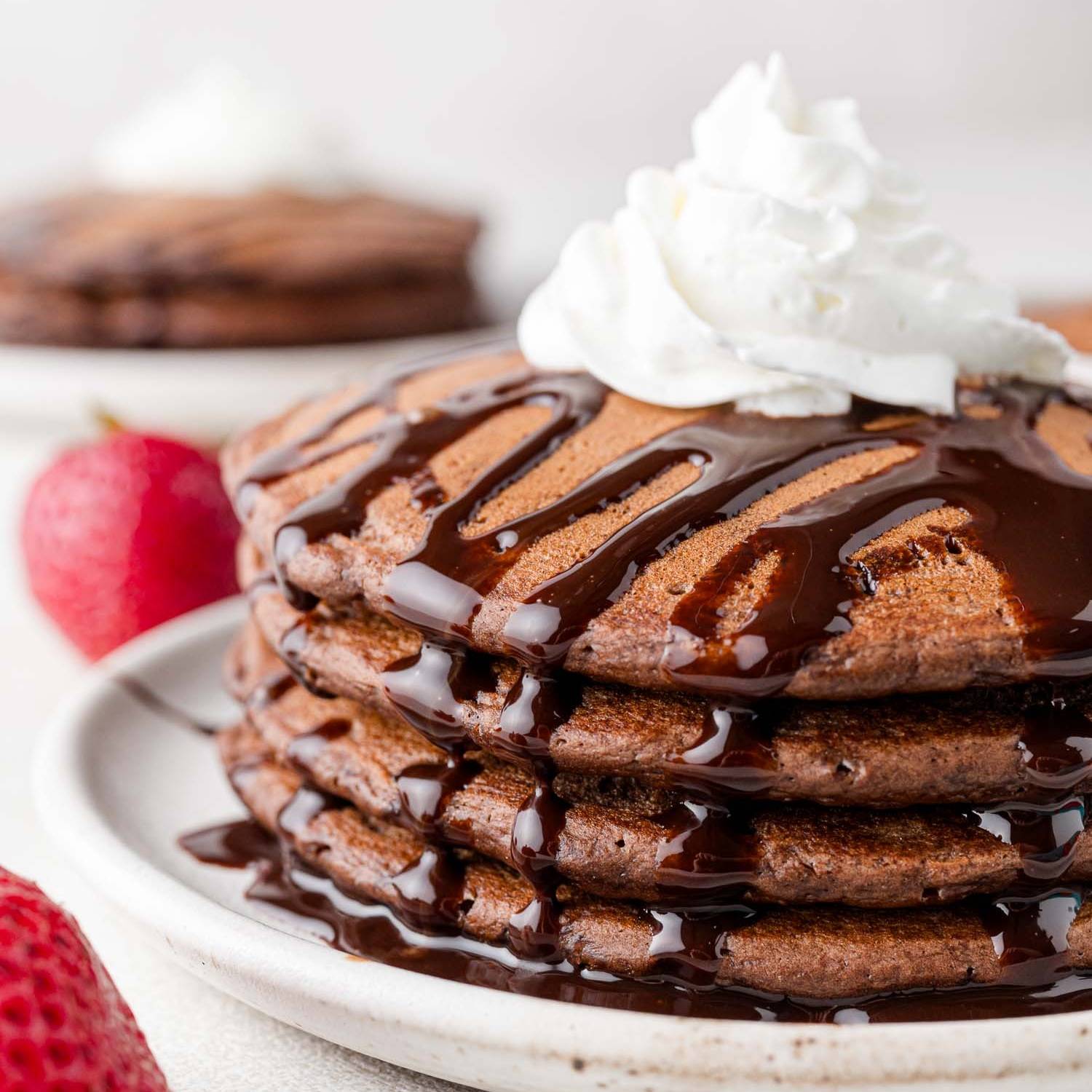 Stack of chocolate pancakes topped with chocolate syrup and whipped cream.
