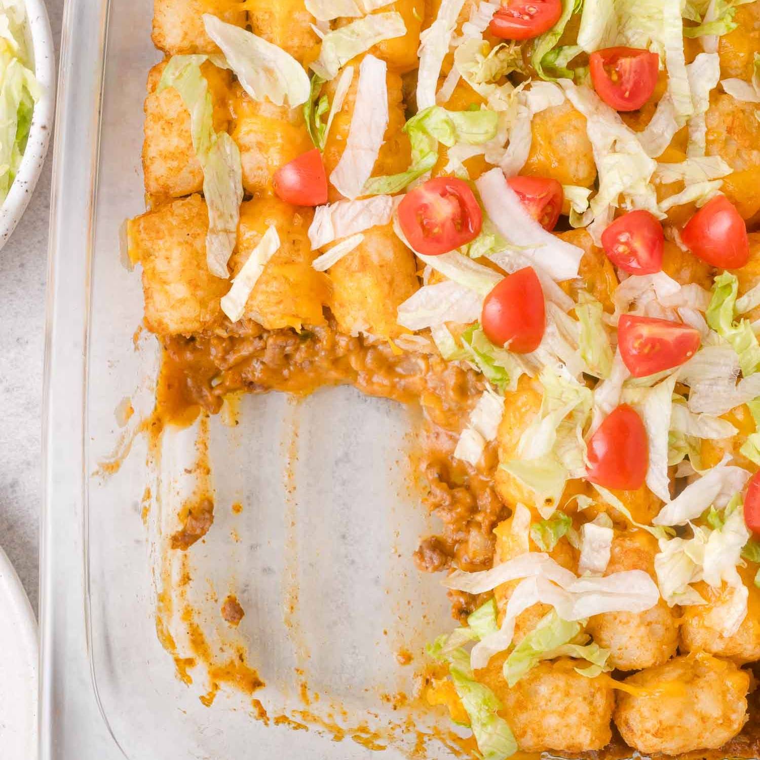 Casserole in a clear dish with a cheesy hamburger layer topped with tater tots, tomatoes, and lettuce.
