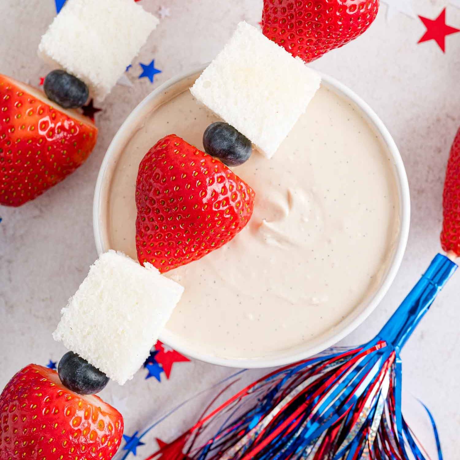 Red, white, and blue fruit kabob resting on a bowl of yogurt dip.