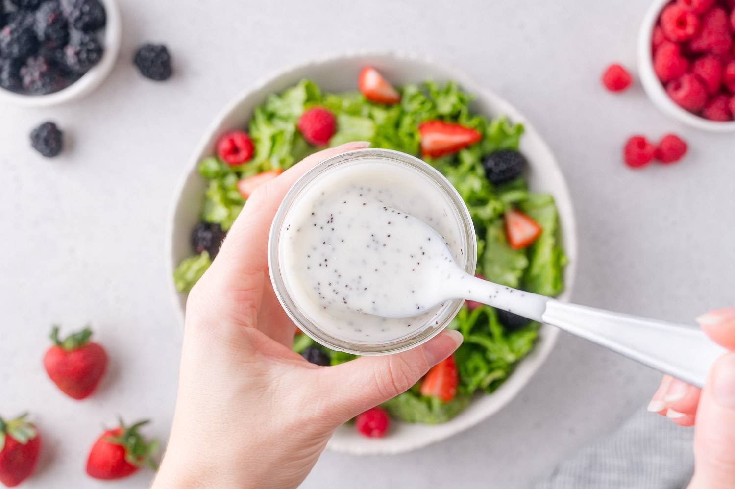 Poppyseed dressing in a jar, and held on a spoon, over a bowl of green salad with berries.