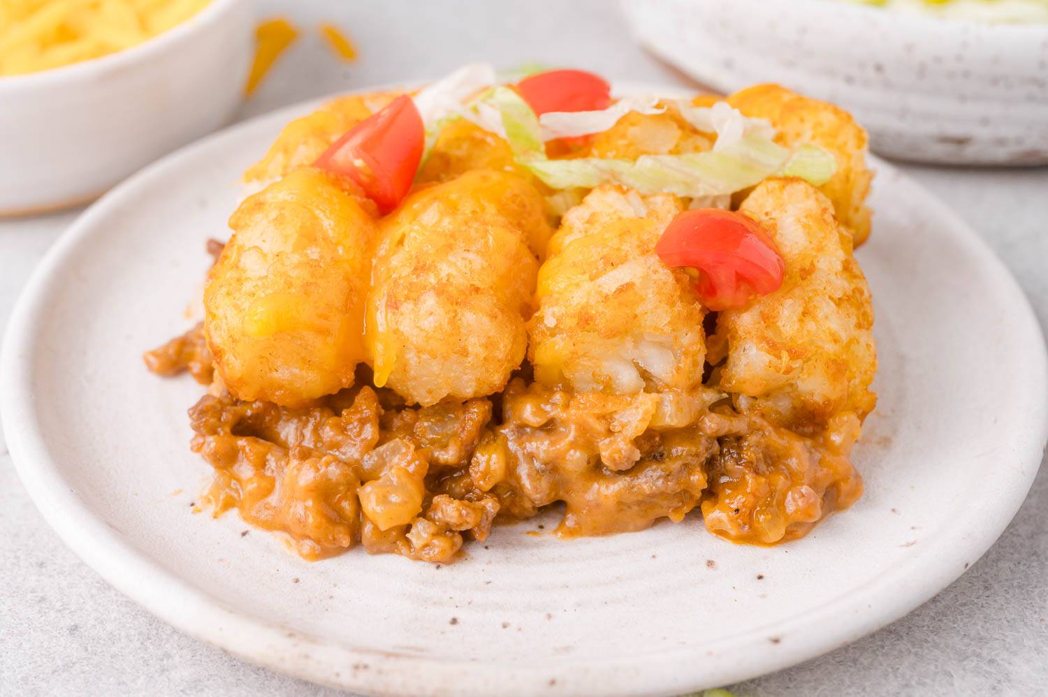 Piece of cheeseburger tater tot casserole, a beef bottom with a tater tot topping, with cheese, lettuce, tomatoes.