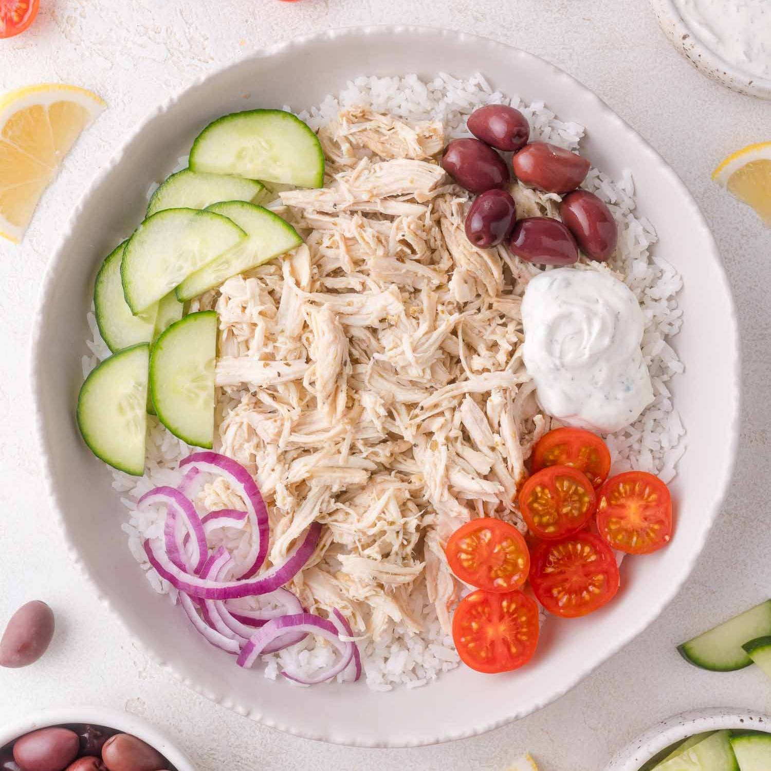 Greek chicken in a bowl with tomatoes, tzatziki, onions, cucumbers, and olives.