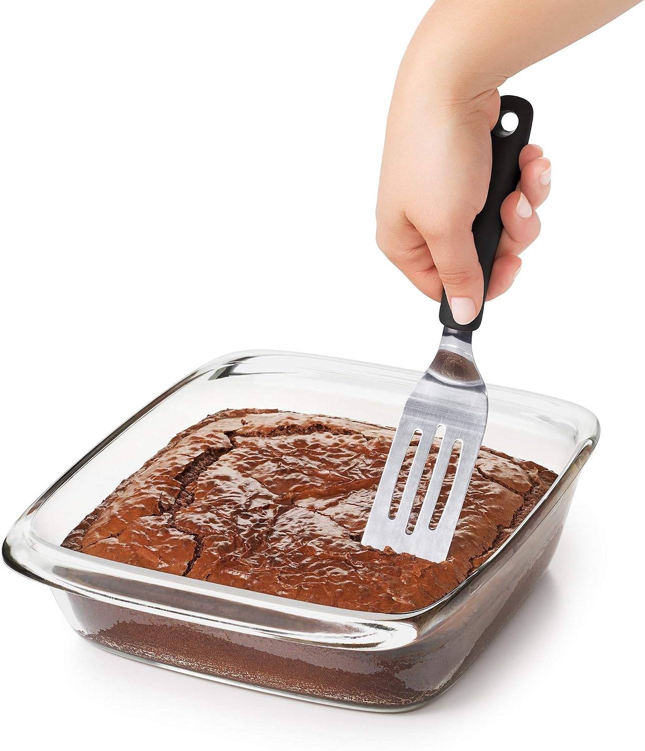 Hand using a small metal spatula to scoop brownies out of a clear glass pan.