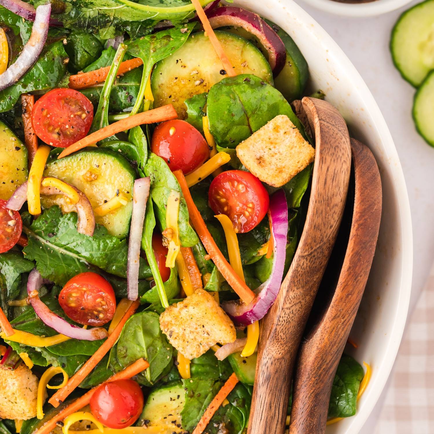 House salad with carrots, onion, tomato, cucumber, and croutons, in a white bowl with wooden serving utensils.