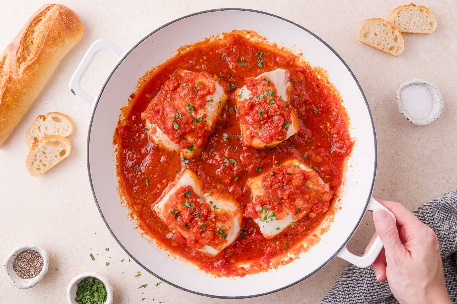 Four pieces of cod in a white pan, with a tomato sauce. A hand holds the pan.
