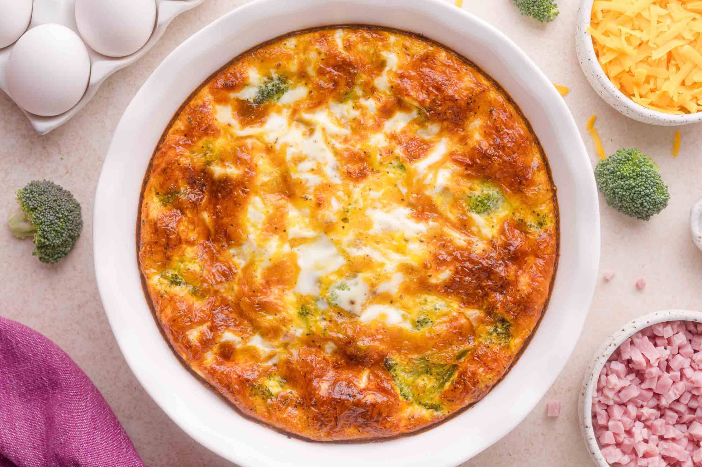 Crustless quiche in a white pie plate, surrounded by broccoli florets, cubed ham, and shredded cheddar.