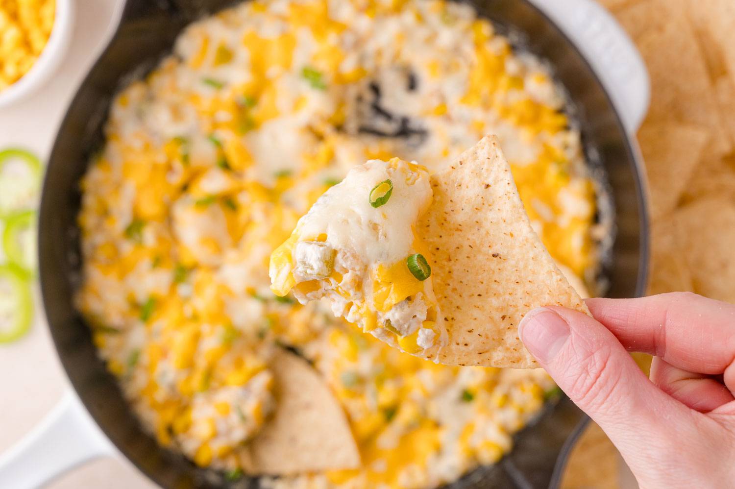 Corn and jalapeno dip in a black skillet, a hand holding a chip with dip over the skillet.