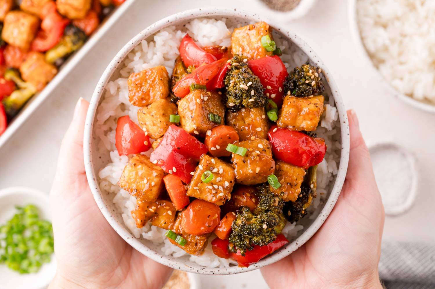 Bowl of General Tso's tofu and vegetables on top of rice, being held with two hands.
