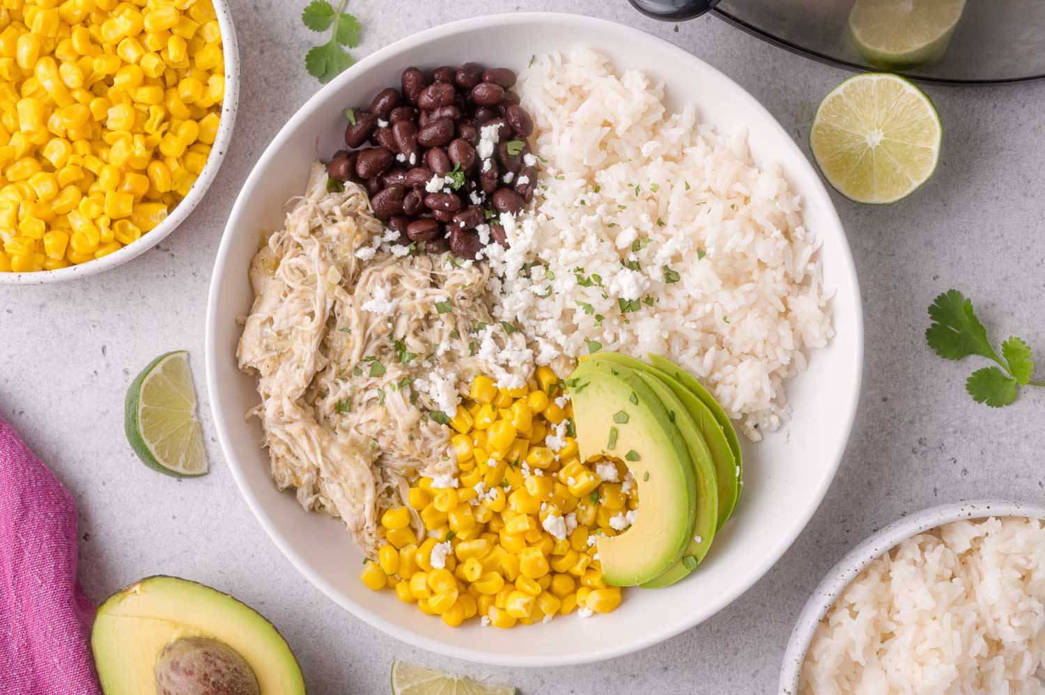 A white bowl with rice, shredded chicken, corn, black beans, and sliced avocado.
