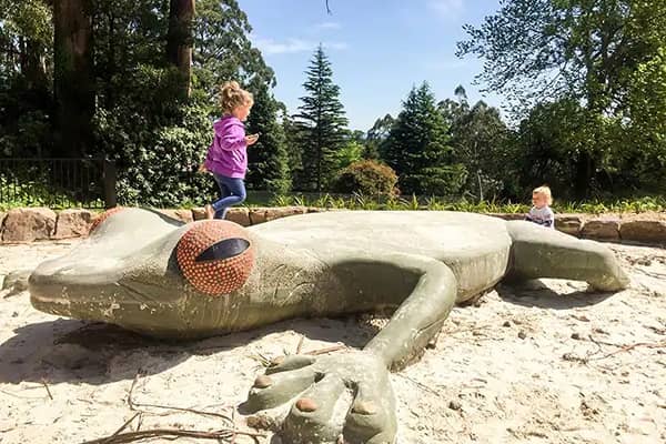 A child in a purple jumper runs past the orange eye of a giant lizard sculpture in a sandy play area, with a toddler sitting nearby and tall trees behind.