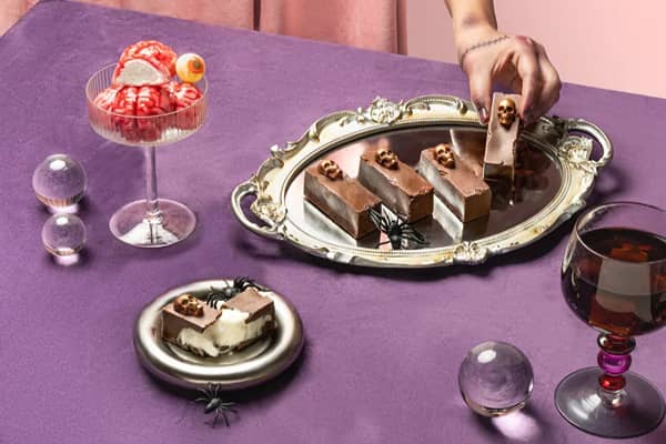 A purple table set with Halloween desserts: a glass of ice cream scoops made to look like brains, chocolate coffin-shaped slices topped with gold skulls on a mirrored tray, a slice of cake, plastic spiders and crystal balls.